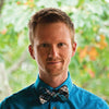 A man with short light brown hair in a bright blue shirt and plaid bow tie stands outside with greenery, smiling slightly while holding The Witch’s Oracle, 2nd Edition by Schiffer Publishing, Ltd.
