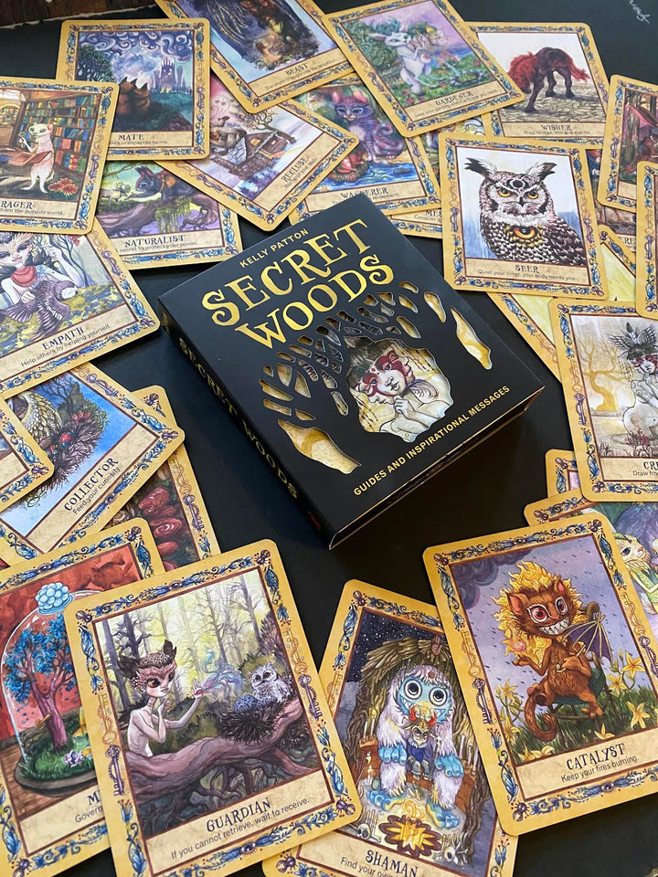 The Secret Woods deck by Schiffer Publishing, Ltd. features illustrated intuition cards with whimsical forest creatures and includes a black and gold guidebook, all displayed on a dark surface.