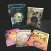 The Hummingbird Wisdom Oracle Cards by Schiffer Publishing, Ltd. are shown in a boxed set with illustrated cards and a guidebook featuring colorful hummingbirds, flowers, and nature scenes arranged on a black surface.