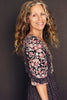 A woman with long, wavy, light brown hair stands sideways, smiling in emotional well-being. She wears a black floral dress. "Listening to Flowers" by Schiffer Publishing, Ltd. is set against a dark, plain background.
