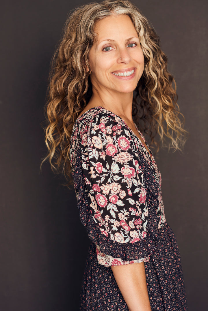 A woman with long, wavy, light brown hair stands sideways, smiling in emotional well-being. She wears a black floral dress. "Listening to Flowers" by Schiffer Publishing, Ltd. is set against a dark, plain background.
