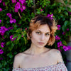 Radiating emotional well-being, a person with short, wavy, light brown hair and a floral top stands before green leaves and magenta flowers, inspired by "Listening to Flowers" from Schiffer Publishing, Ltd.