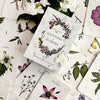The book "Listening to Flowers" by Schiffer Publishing, Ltd. rests on illustrated flower cards featuring botanical drawings and Bach flower essence insights, surrounded by white blooms and greenery.