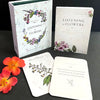The Listening to Flowers tarot-style card deck by Schiffer Publishing, Ltd. is shown with its box, guidebook, and two illustrated cards on a black surface, alongside two orange flowers.