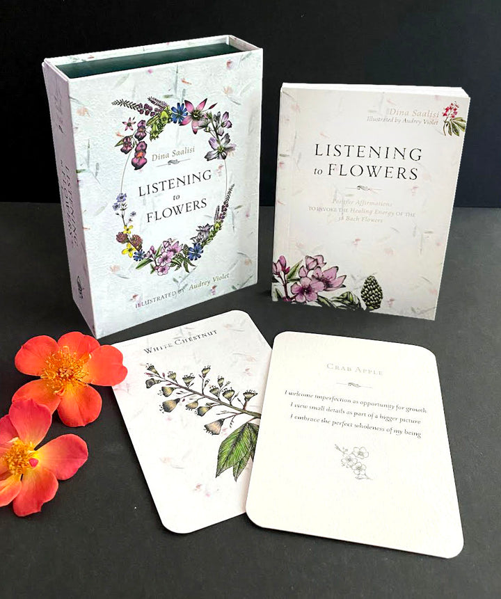 The Listening to Flowers tarot-style card deck by Schiffer Publishing, Ltd. is shown with its box, guidebook, and two illustrated cards on a black surface, alongside two orange flowers.