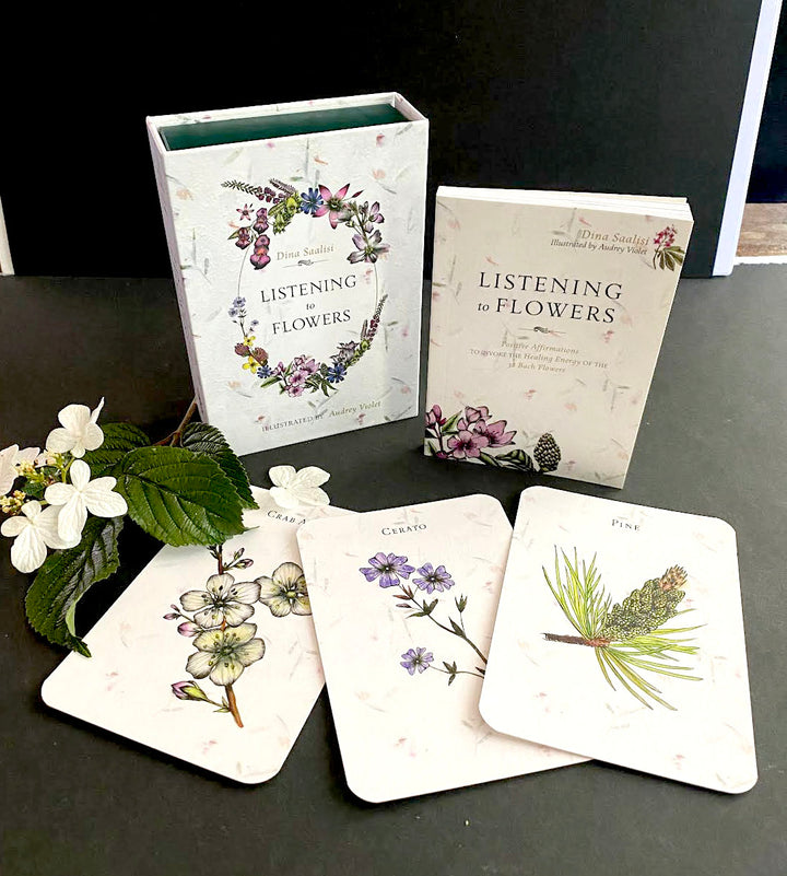The Listening to Flowers card set from Schiffer Publishing, Ltd. is shown on a black surface with three illustrated cards—camellia, cyani, and pine—and a sprig of white flowers beside it, highlighting themes of emotional well-being.