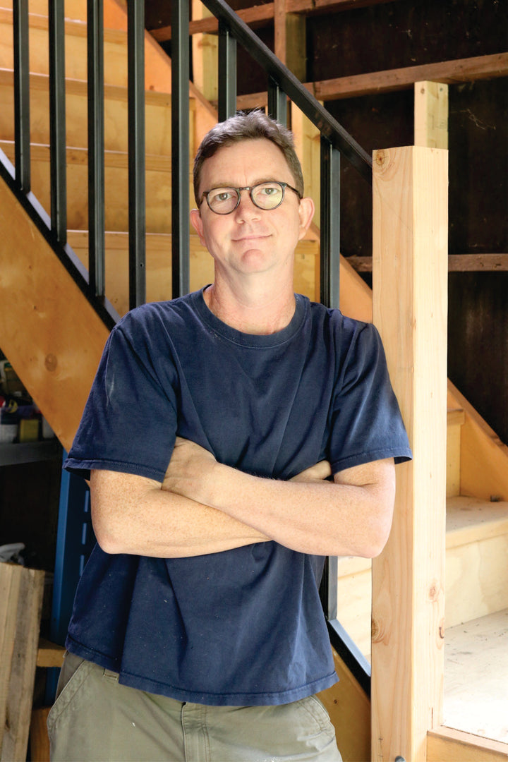 A man with short brown hair and glasses, wearing a navy blue t-shirt and green pants, stands in a workshop by wooden stairs, ready to share his Pinball expertise from Schiffer Publishing, Ltd.