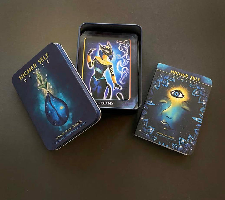 The Higher Self Oracle by Schiffer Publishing, Ltd. is shown with its tin box, a card featuring a mystical cat, and a guidebook with an abstract face—ideal spiritual guidance cards for seekers.