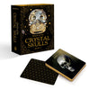 A box labeled "Oracle of the Crystal Skulls" by Schiffer Publishing, Ltd. stands upright beside its oracle deck; one card features a skull and "Jade," suggesting spiritual growth, while another displays a black-and-gold geometric design.