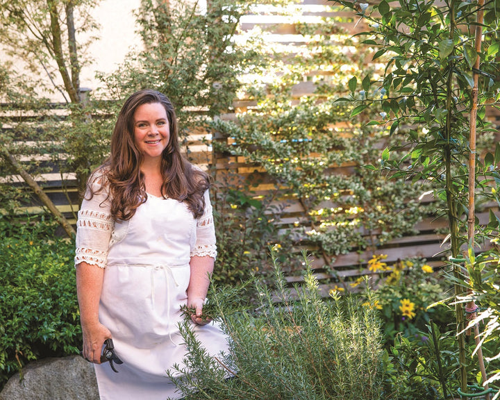 A woman with long brown hair smiles in a lush garden, pruning shears in hand—perfect inspiration for homemade recipes using garden ingredients, as featured in Schiffer Publishing, Ltd.'s Big Book of Botanical Crafts.