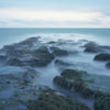 Village Beaches" by Schiffer Publishing, Ltd. captures a serene, surreal long exposure of a mossy rocky jetty on East Hampton beaches at sunrise or sunset, with misty water and a calm ocean beneath cloudy skies.
