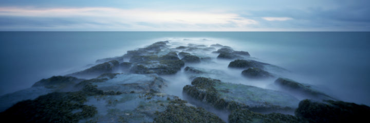 Village Beaches" by Schiffer Publishing, Ltd. captures a serene, surreal long exposure of a mossy rocky jetty on East Hampton beaches at sunrise or sunset, with misty water and a calm ocean beneath cloudy skies.