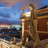 A stone statue overlooks a Northern Italian city at dusk, with glowing buildings and a distant harbor. Explore hidden gems with "Secret Cities of Italy" by Schiffer Publishing, Ltd.—your guide to discovering Italy’s lesser-known destinations.