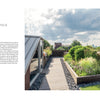 Repose in the Metropolis by Schiffer Publishing, Ltd. captures a modern rooftop garden with wooden walkways, raised planters of green shrubs and grasses, and city skyline views beneath a partly cloudy sky.