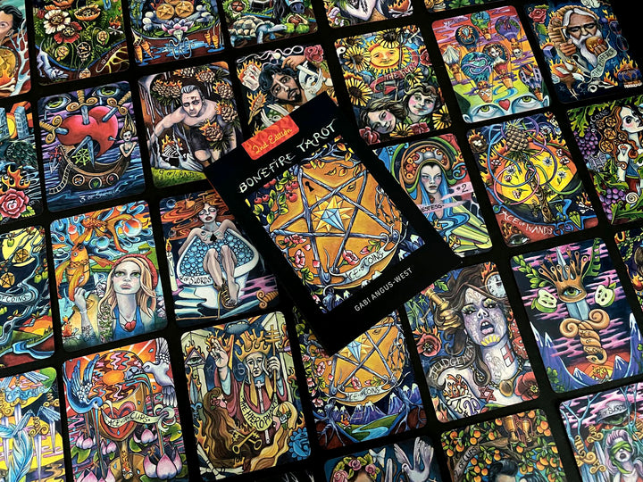 A vibrant display of Bonefire Tarot cards by Schiffer Publishing, Ltd. features colorful, hand-painted illustrations. The deck’s box displays a golden pentacle, resting atop scattered cards filled with expressive, mystical imagery.