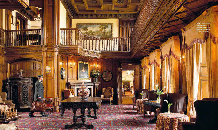 In "At Home with Royalty" by Schiffer Publishing, Ltd., experience a grand castle room with wood paneling, gold-curtained windows, antique furniture, and a suited man near the fireplace below a balcony adorned with artwork.