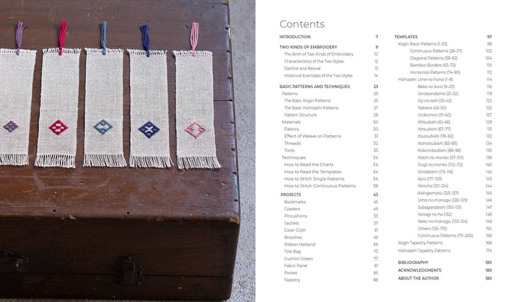 Five geometric woven bookmarks in red, blue, and purple—including hishizashi and Kogin styles—are arranged on a wooden chest beside the contents page of "Counted Sashiko Embroidery" by Schiffer Publishing, Ltd.