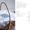 A wicker basket holds Schiffer Publishing, Ltd.'s Counted Sashiko Embroidery—a white cloth with a blue geometric hishizashi-inspired pattern. Nearby, a diagram shows the cloth's dimensions, pattern placement, and details about the cover cloth.