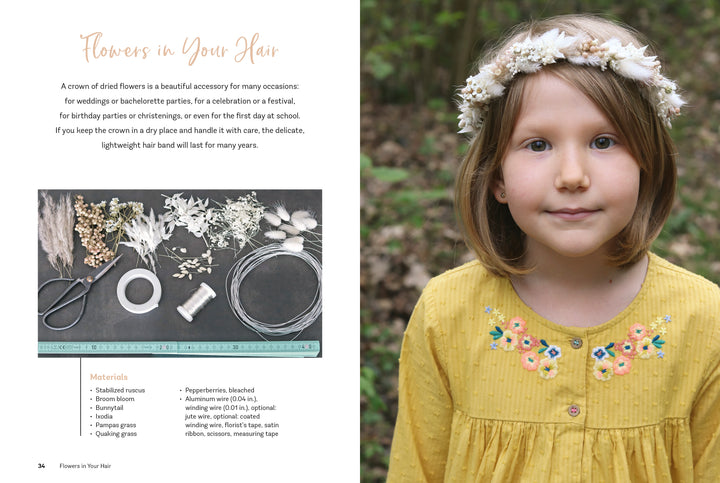 A young girl with short brown hair wears a handmade white floral crown from dried flowers and a yellow dress with blue embroidery. Next to her, Dried Flower Love by Schiffer Publishing, Ltd. displays dried flower craft materials outdoors on a table.