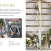 The "Dried Flower Love" wreath by Schiffer Publishing, Ltd. features dried flowers, greenery, twine, and quail eggs. It hangs on a white wall beside a workspace filled with crafting tools and botanicals—perfect for your next home décor project.
