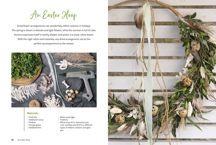 The "Dried Flower Love" wreath by Schiffer Publishing, Ltd. features dried flowers, greenery, twine, and quail eggs. It hangs on a white wall beside a workspace filled with crafting tools and botanicals—perfect for your next home décor project.