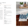 Two Nordic Hands towels by Schiffer Publishing, Ltd.—white with blue Scandinavian-inspired geometric patterns—hang on a wooden kitchen drawer near a white sink. In the background: a plant, candle, and glass jars on the counter.