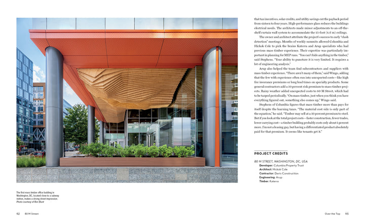 The entrance, inspired by "Innovations in Mass Timber" from Schiffer Publishing, Ltd., features geometric timber ceilings, expansive glass, and bold red-orange columns, showcasing the environmental benefits of modern commercial architecture amid urban greenery.