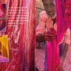A man dyes yarn bright pink outdoors, his stained hands and vivid threads inspired by Rajasthan. Colorful woven fabric appears at the right. The scene is from "The Pigment Trail" by Schiffer Publishing, Ltd., inspired by Indian decorative arts.
