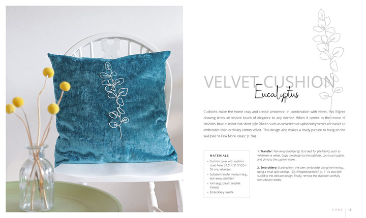 A blue velvet "Stitch with One Line" cushion by Schiffer Publishing, Ltd., featuring a white one-line eucalyptus embroidery, rests on a white chair beside a vase of yellow billy balls. Text to the right details the cushion and its materials.