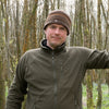 A man in a brown striped beanie and olive zip-up jacket stands in a forest, smiling at the camera—ready to share outdoor tips from Core Bushcraft by Schiffer Publishing, Ltd.