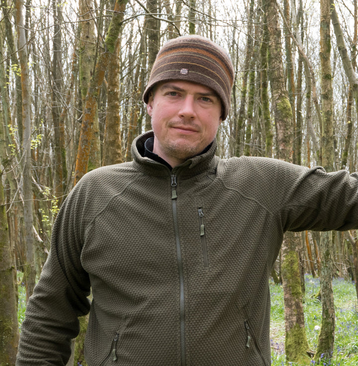 A man in a brown striped beanie and olive zip-up jacket stands in a forest, smiling at the camera—ready to share outdoor tips from Core Bushcraft by Schiffer Publishing, Ltd.