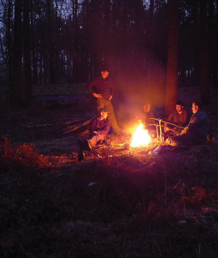 Four people sit and one stands around a glowing campfire at dusk, sharing stories and skills featured in Core Bushcraft by Schiffer Publishing, Ltd. Their faces are lit by firelight amid tall trees, creating a peaceful outdoor atmosphere.