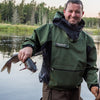 A man in a green jacket smiles by a river, holding a fishing rod and catfish—highlighting his wilderness skills inspired by Core Bushcraft from Schiffer Publishing, Ltd., against a scenic backdrop of trees and water.