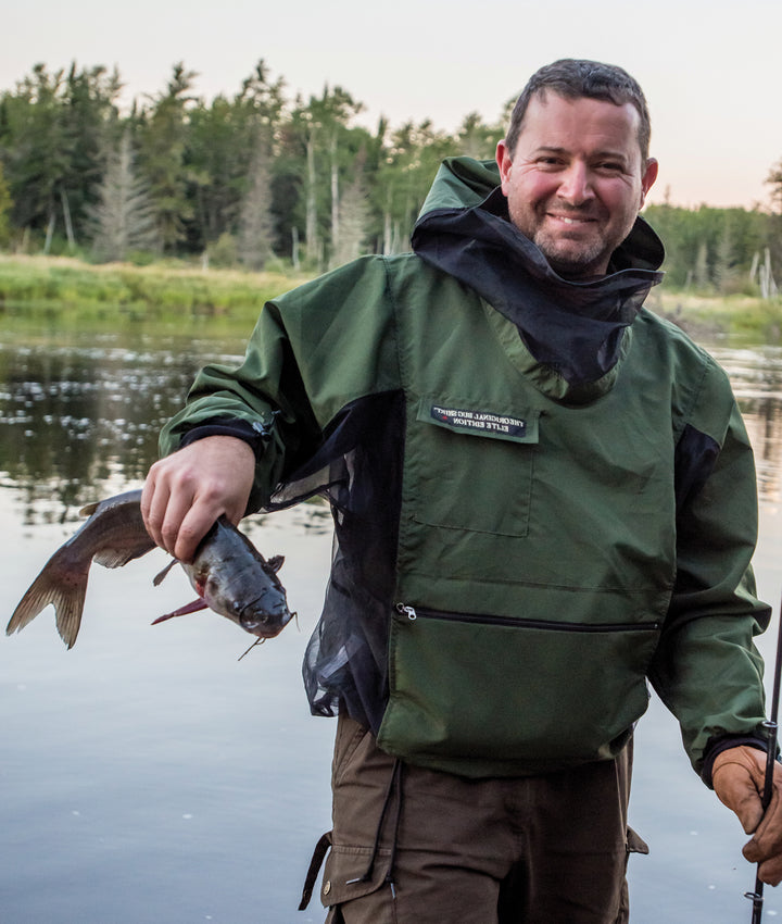 A man in a green jacket smiles by a river, holding a fishing rod and catfish—highlighting his wilderness skills inspired by Core Bushcraft from Schiffer Publishing, Ltd., against a scenic backdrop of trees and water.