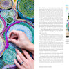 A woman smiles at a table with colorful, circular crocheted art from "Threads of Treasure" by Schiffer Publishing, Ltd. On the left, her hand embroiders vibrant patterns, inspiring today’s modern crafters.