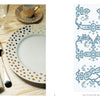 A table set with gold-dotted white plates, embroidered burlap napkin, silverware, wine glass, and candle holder sits beside Schiffer Publishing, Ltd.'s Vintage French Needlework featuring blue monogram patterns on graph paper.