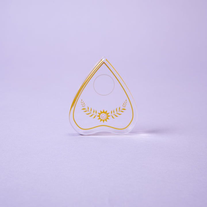 The Crystal Spirit Talking Board and Guidebook by Schiffer Publishing, Ltd. is a transparent, heart-shaped board with a circular top hole and gold design, standing upright on a light purple surface, radiating sacred crystal energy.