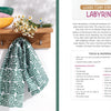 A green and white tea towel, crafted with the Fabric Stamping technique from Schiffer Publishing, Ltd., hangs on a wooden rack beside stacked bowls and daisies. Foam stamp printing instructions appear to the right.