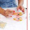 A person uses Schiffer Publishing, Ltd.’s Fabric Stamping to make pink and yellow prints with a clear flower stamp on white fabric, surrounded by paint bottles and stamping tools. Foam-stamping project text is shown on the right.