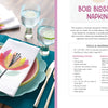 A fabric-stamped flower napkin from Schiffer Publishing, Ltd.'s Fabric Stamping is shown on a white plate with a blue charger. Nearby are a napkin and stamp, plus the title Bold Blossom Napkins and a list of tools and materials.