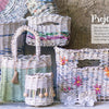 A display of Weaving Paper baskets by Schiffer Publishing, Ltd.—upcycled waste paper baskets in assorted shapes and sizes, some with colorful accents and flowers, arranged on light wood with a large patterned basket in the background.