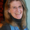 A smiling woman with long light brown hair and glasses wears a dark turtleneck and a blue-patterned scarf, standing by a light wall. She holds "Swatch Critters from the Pin Loom" by Schiffer Publishing, Ltd.
