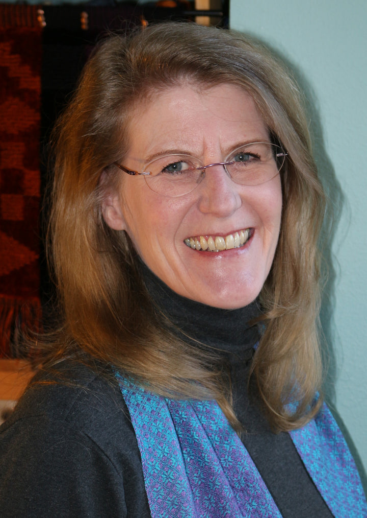 A smiling woman with long light brown hair and glasses wears a dark turtleneck and a blue-patterned scarf, standing by a light wall. She holds "Swatch Critters from the Pin Loom" by Schiffer Publishing, Ltd.