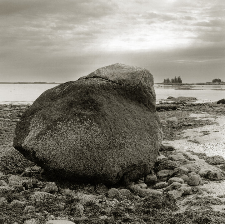 A large, weathered boulder sits on a rocky shore beneath cloudy skies in "The Maine Coast" by Schiffer Publishing, Ltd., evoking tranquil moods with distant trees and calm water in the background.