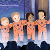Four children in orange astronaut suits stand arm-in-arm, waving for cameras by a rocket launchpad at night in "Reaching for the Stars" from Schiffer Publishing, Ltd.—a picture book capturing the excitement of NASA Artemis missions and space exploration.