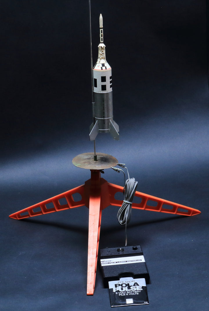 A Schiffer Publishing, Ltd. Model Rocketry kit is set up on a red three-legged launch pad, with a corded control unit attached; the dramatic dark background highlights the rocket and setup, recalling the golden age of space flight.