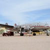 A Lockheed Constellation by Schiffer Publishing, Ltd. is displayed on a sandy lot with other planes nearby, a red support vehicle near its nose, and yellow chocks around its landing gear.