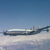 A Lockheed Constellation airplane soars above the clouds in a clear blue sky. This classic four-engine aircraft, featured by Schiffer Publishing, Ltd., displays the vintage Lufthansa logo on its tail.