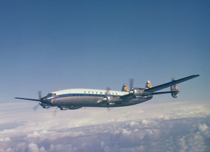 A Lockheed Constellation airplane soars above the clouds in a clear blue sky. This classic four-engine aircraft, featured by Schiffer Publishing, Ltd., displays the vintage Lufthansa logo on its tail.