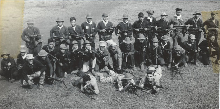A black-and-white photo from "The Fighting Fathers" (Schiffer Publishing, Ltd.) shows soldiers—some linked to CIA South Vietnam ops—posing outdoors with rifles or submachine guns in mixed uniforms and hats, standing, kneeling, or prone on the grass.
